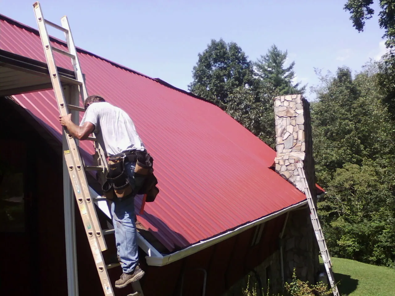 Roofer on a ladder beside a red metal roof and stone chimney — Soffit & Fascia Repair by Roofing Brothers in Hyde Park